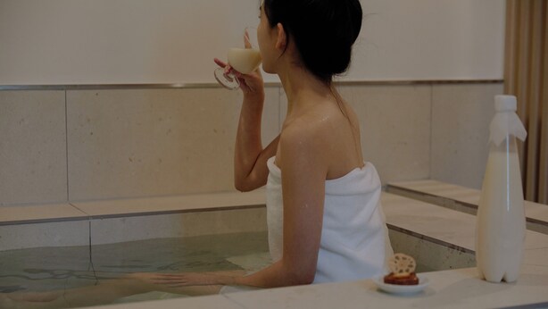 Eine Person liegt in einer Badewanne, umhüllt von einem Handtuch, mit einer Lotionflasche und einer kleinen Schale mit Snacks.