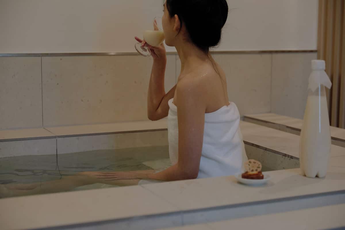 Eine Person liegt in einer Badewanne, umhüllt von einem Handtuch, mit einer Lotionflasche und einer kleinen Schale mit Snacks.