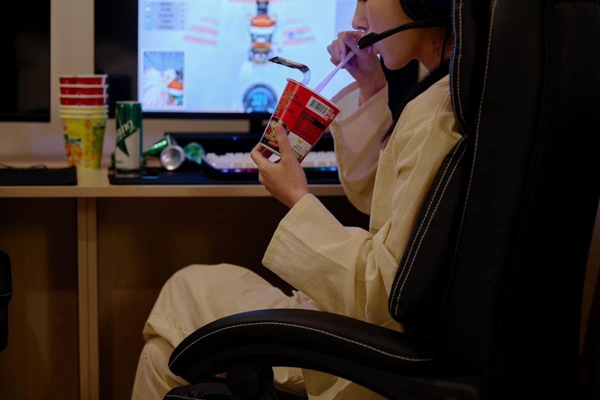 Eine Person sitzt in einem Sessel, hält eine Tasse und eine Dose, und im Hintergrund ist ein Bildschirm mit einem Spiel zu sehen.