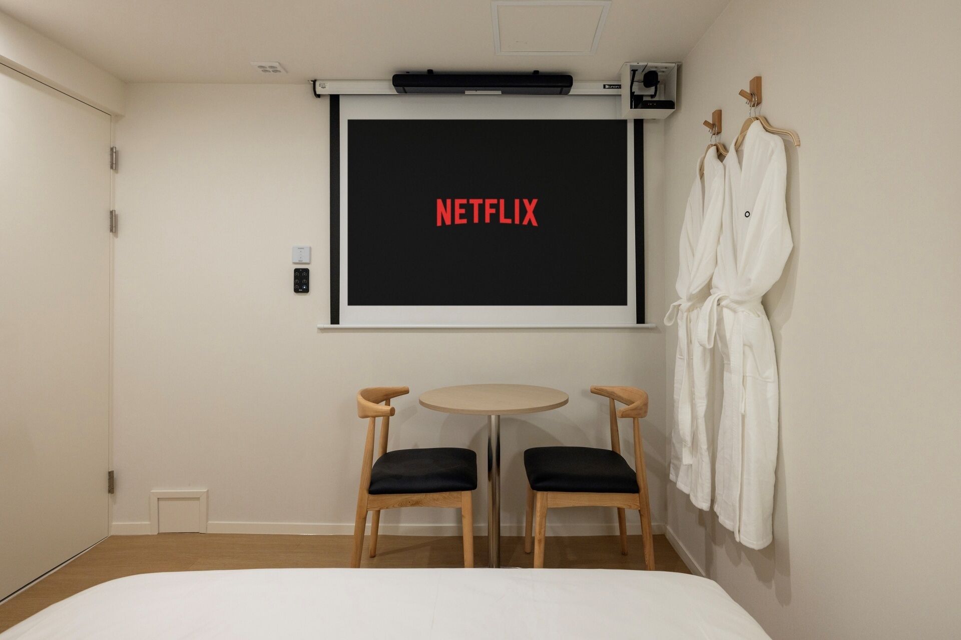 A hotel room with a bed, two chairs, a small table, a TV screen displaying 'NETFLIX', and a wall-mounted robe rack.