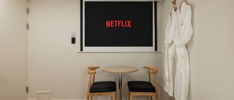 A hotel room with a bed, two chairs, a small table, a TV screen displaying 'NETFLIX', and a wall-mounted robe rack.