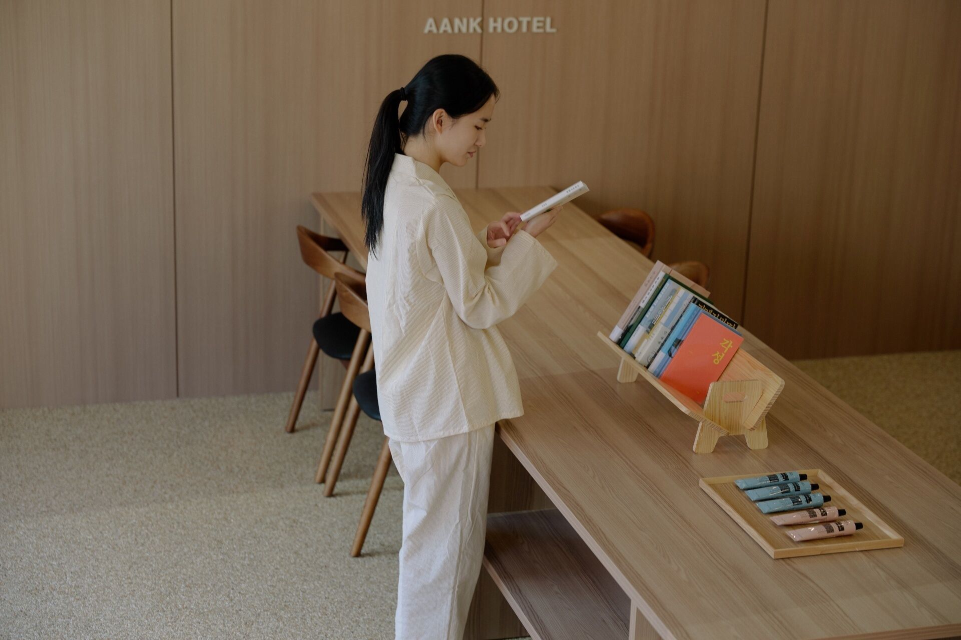 A person reading a book in a room with a wooden wall, a wooden table with books and toiletries, and a bench.