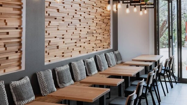 A modern restaurant with a long bench seating area, wooden wall panels, hanging pendant lights, and potted plants.