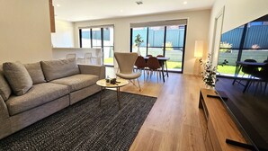 A modern living room with a sofa, a coffee table, and a dining area with chairs and a table. There is a large window providing a view of the outdoors.