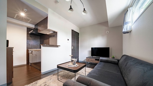 Un salon moderne avec un canapé gris foncé, un téléviseur à écran plat et un coin cuisine équipé d’une cuisinière et de placards.