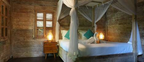 A wooden four-poster bed with a canopy, a nightstand with a lamp, and a window with shutters.