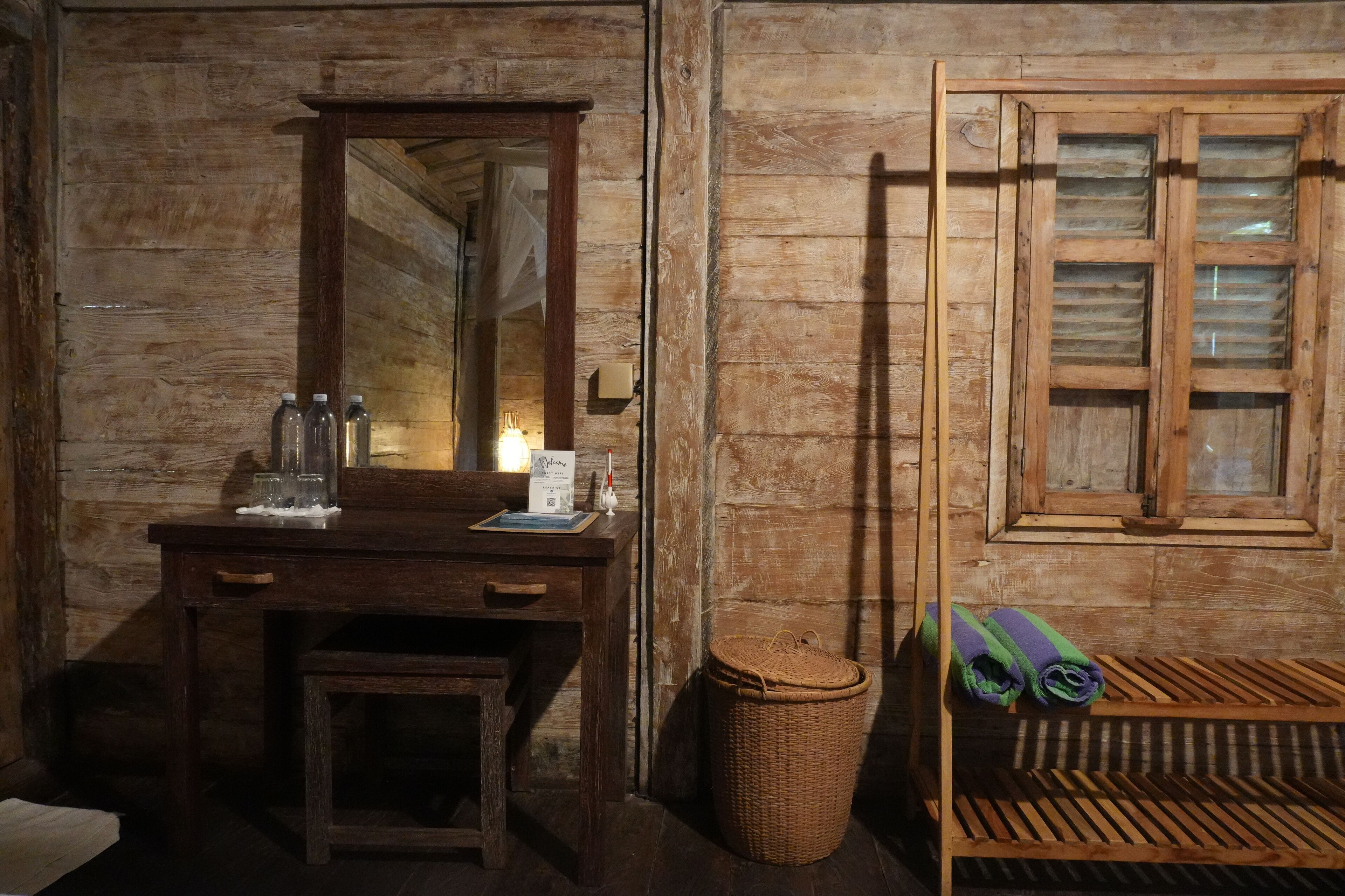 A rustic wooden room with a wooden desk, a mirror, a wicker basket, and a window with shutters.
