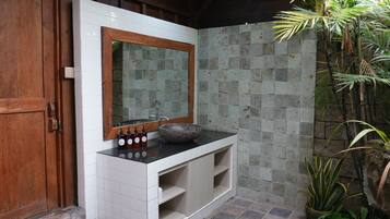 Un baño con puerta de madera, pared de azulejos blancos, pared de piedra, encimera negra, tocador de madera, espejo grande y una planta en maceta.
