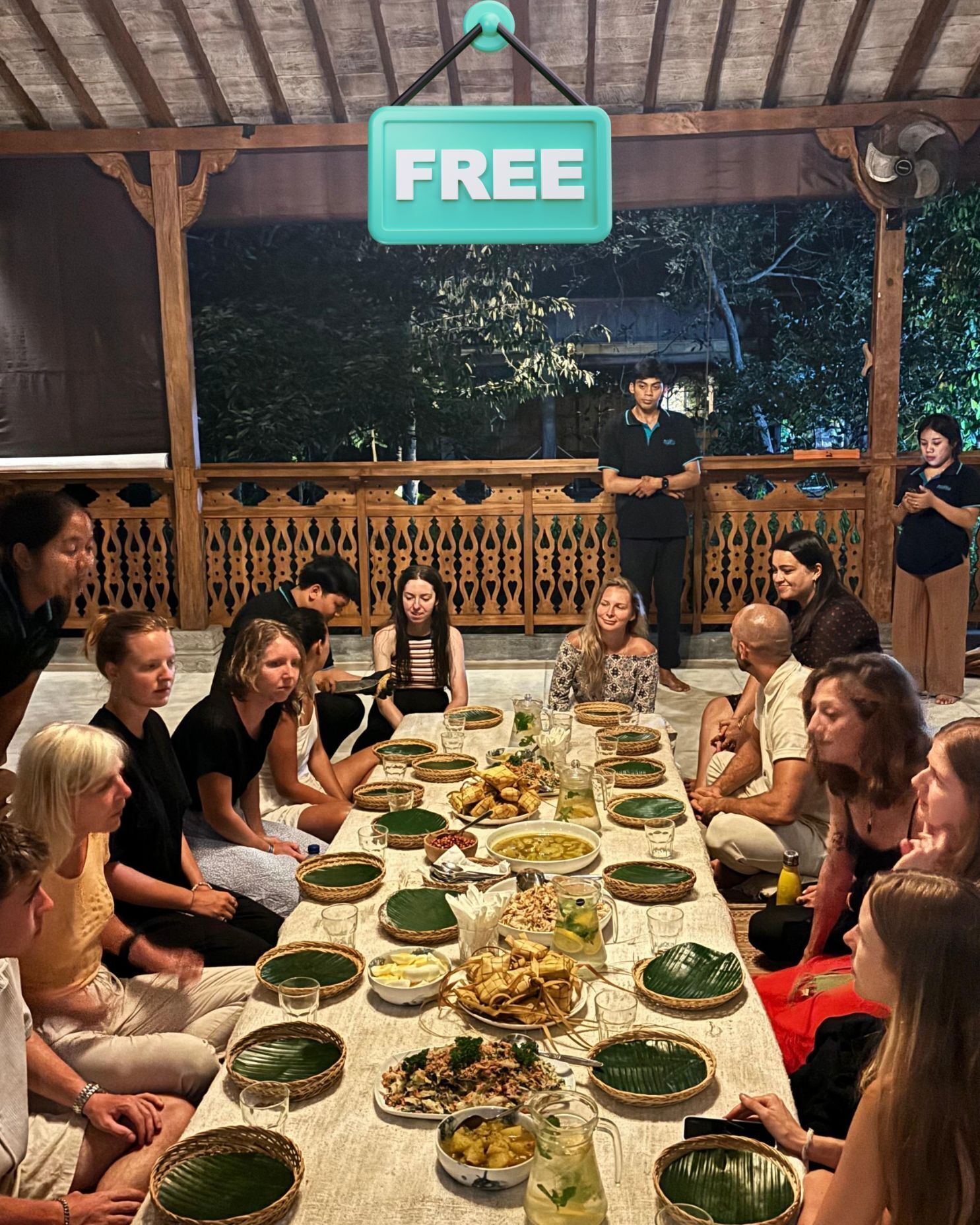 A group of people seated around a long table with various dishes, under a wooden structure with a 'FREE' sign above.