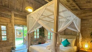 A wooden four-poster canopy bed with a white bedspread and teal pillows, a nightstand with a lamp, and a view of the outdoors through a window. - Nunu Bali Eco Friendly Retreat (Tabanan)