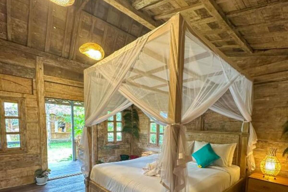 Un letto a baldacchino in legno con tende bianche e cuscini color petrolio, un comodino con una lampada e vista sull'esterno attraverso una finestra.