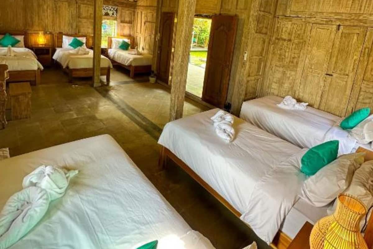 Una stanza rustica in legno con quattro letti, ognuno con lenzuola bianche e cuscini verdi. Sono presenti luci a sospensione e una lampada intrecciata su un tavolo.
