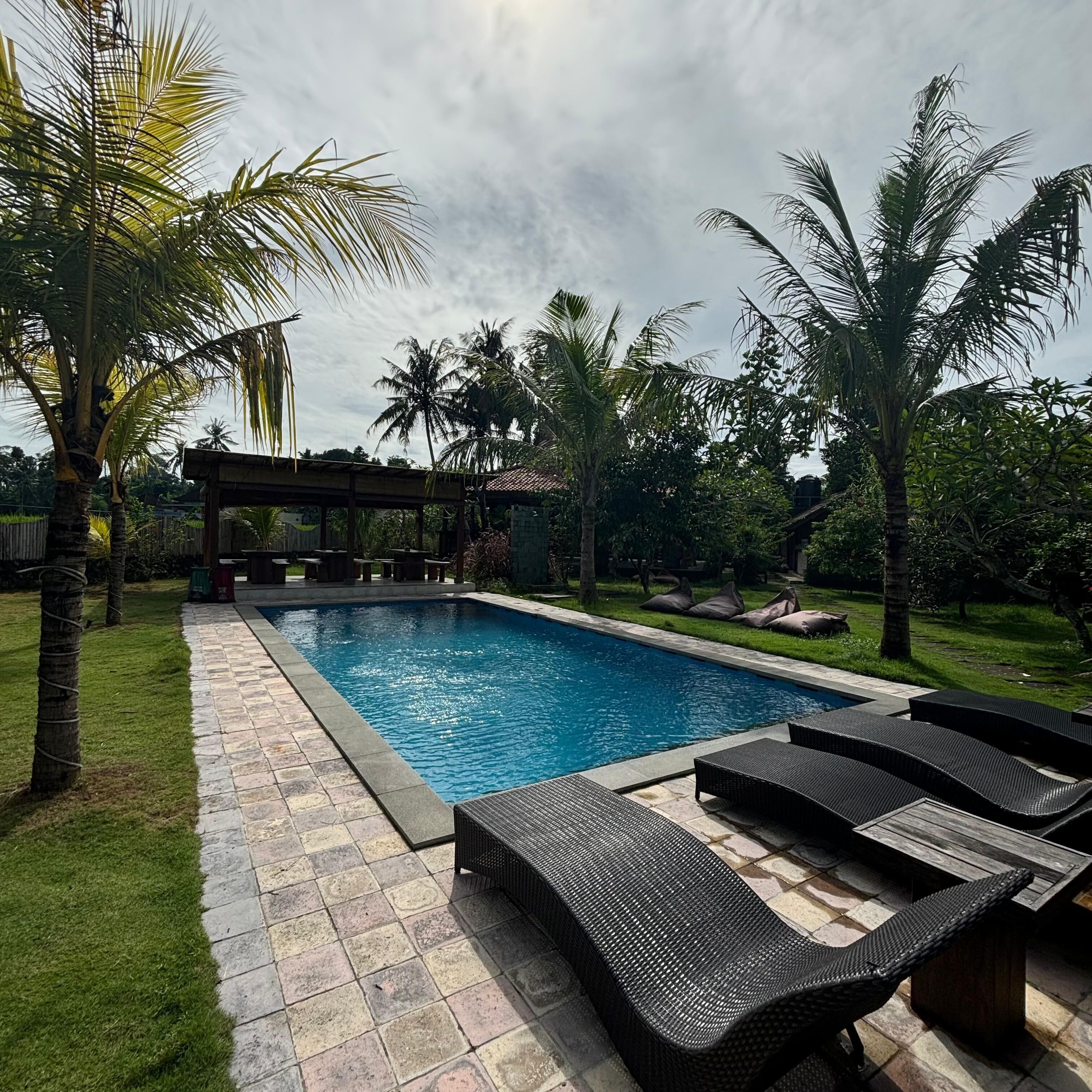 A swimming pool with lounge chairs, surrounded by palm trees and a covered seating area.