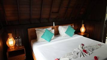 Techo de madera, cama con ropa de cama blanca, almohadas color turquesa, mesita de noche con lámpara y un cajón a un lado de la cama.