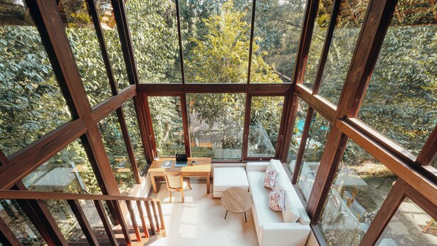A modern interior with a wooden staircase, a white sofa, a wooden table, and a view of a lush garden through large windows. - Nunu Bali Eco Friendly Retreat (Tabanan)