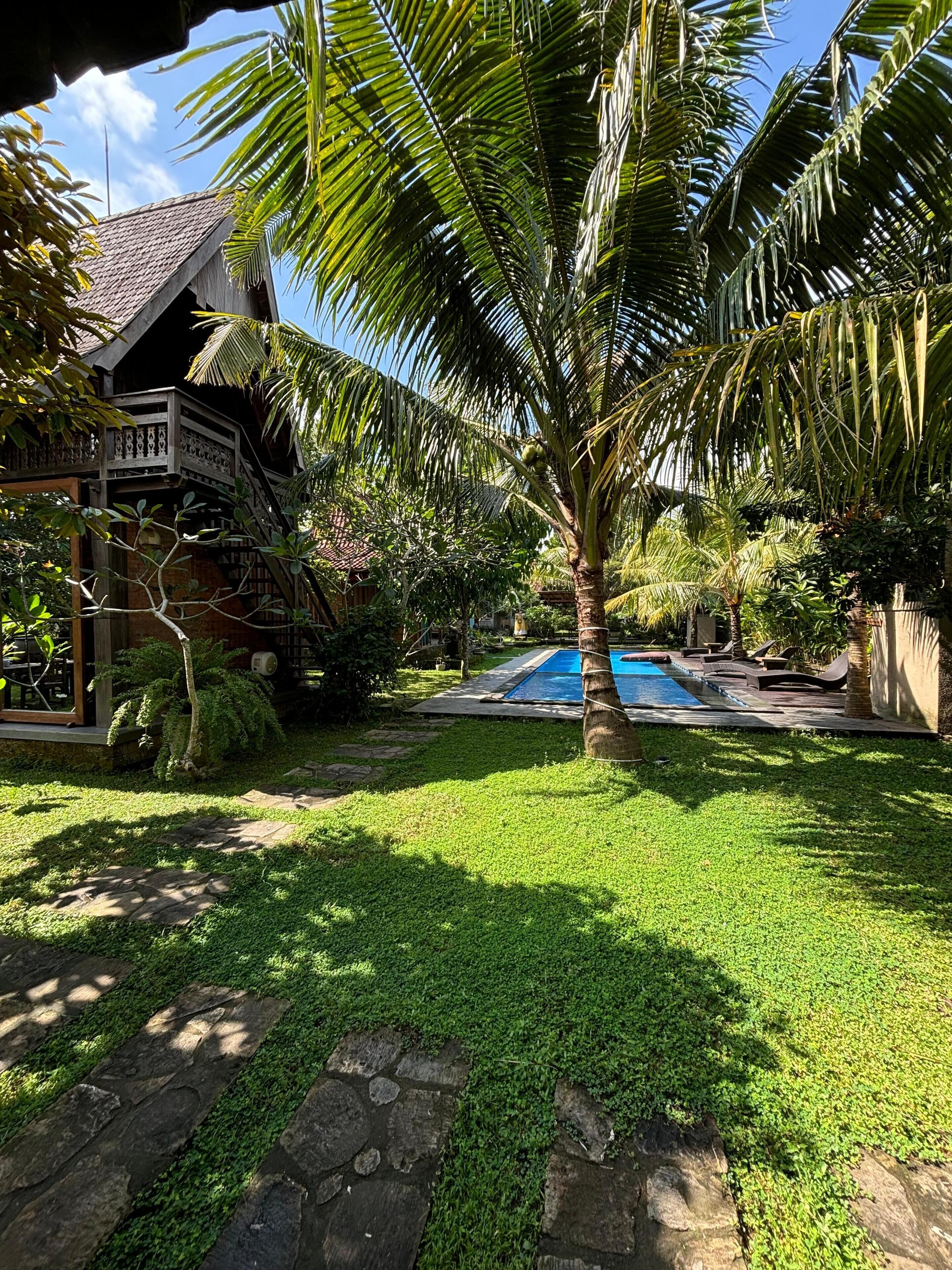 A tropical garden with a swimming pool, stone pathway, and a wooden house with a balcony.