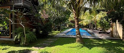A tropical garden with a swimming pool, stone pathway, and a wooden house with a balcony.
