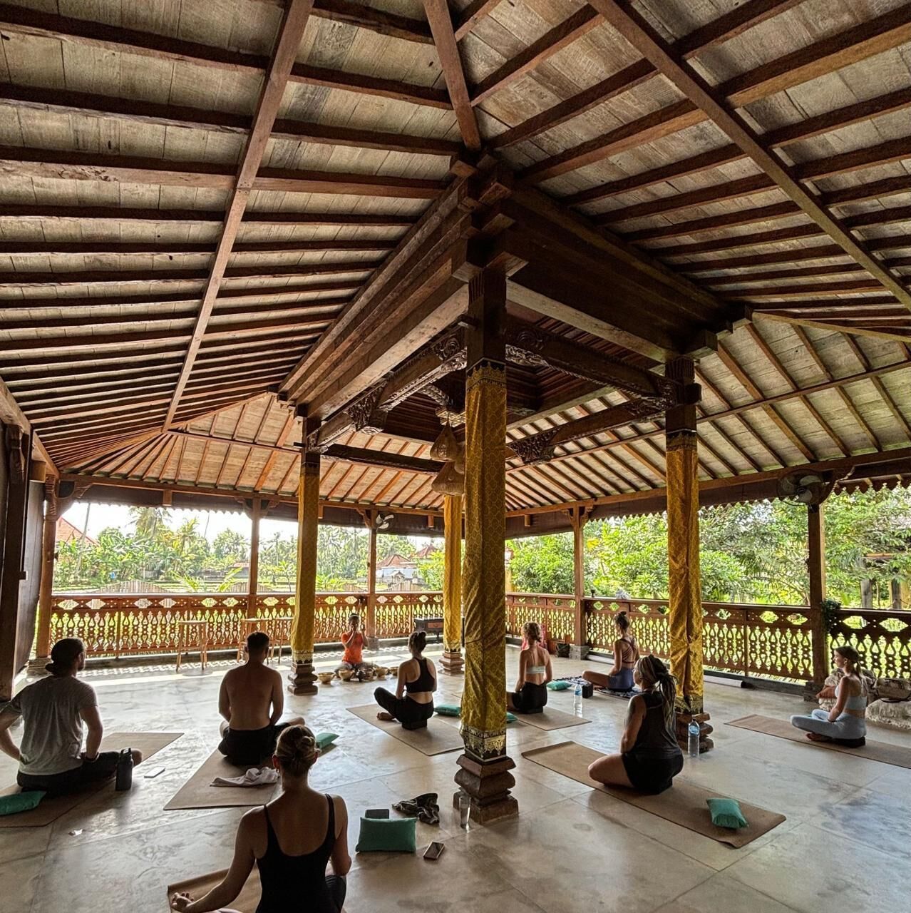 Espacio al aire libre techado para yoga, con vigas y columnas de madera, personas practicando yoga y vistas a la vegetación.