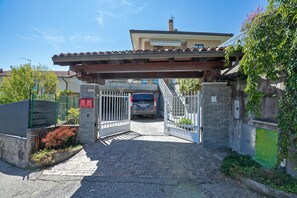 A gated entrance to a property with a covered driveway, a vehicle parked inside, and a building with a tiled roof in the background. - A Due Passi Dal Bosco: Hillside Apartment with Garden&BBQ, Laveno Mombello, Italy (Laveno Mombello)