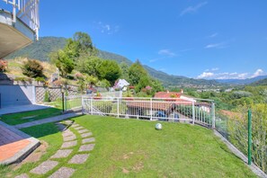 A well-maintained garden with a paved pathway, a white fence, and a view of a hillside town. - A Due Passi Dal Bosco: Hillside Apartment with Garden&BBQ, Laveno Mombello, Italy (Laveno Mombello)