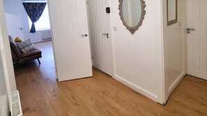 A hallway with white walls, wooden flooring, and a door leading to a room with a window and curtains. - Forge Lane Cozy Apartment (Gillingham)