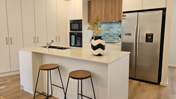 A modern kitchen with white cabinetry, a stainless steel refrigerator, a microwave, and a sink. There are two wooden stools at the island.