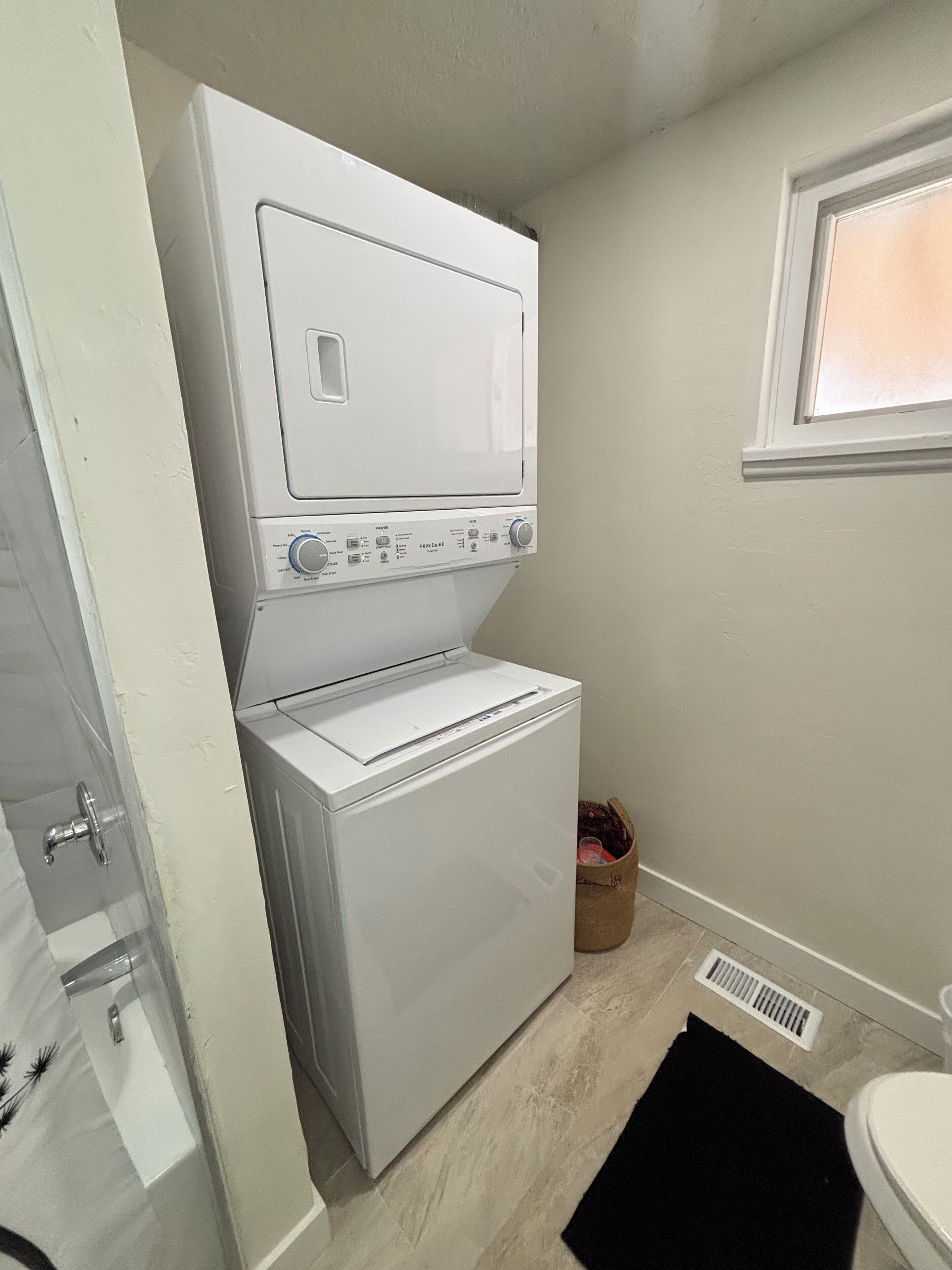 Un lavadero con lavadora y secadora apiladas, una ventana y una puerta que da a un baño.