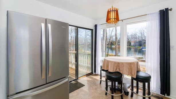 A modern kitchen with a stainless steel refrigerator, a dining area with a table and two stools, and a large window with a view of a lake and deck. - Lakefront Access - Hour from DC - Kayaks Provided (Locust Grove)