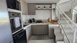 A compact kitchen with a built-in oven, microwave, and dishwasher. There is a small island with a sink and a potted plant. A staircase with a railing leads to an upper level. - Beautiful beachfront penthouse, right on the sand, with a private barbecue area. (Salvador)