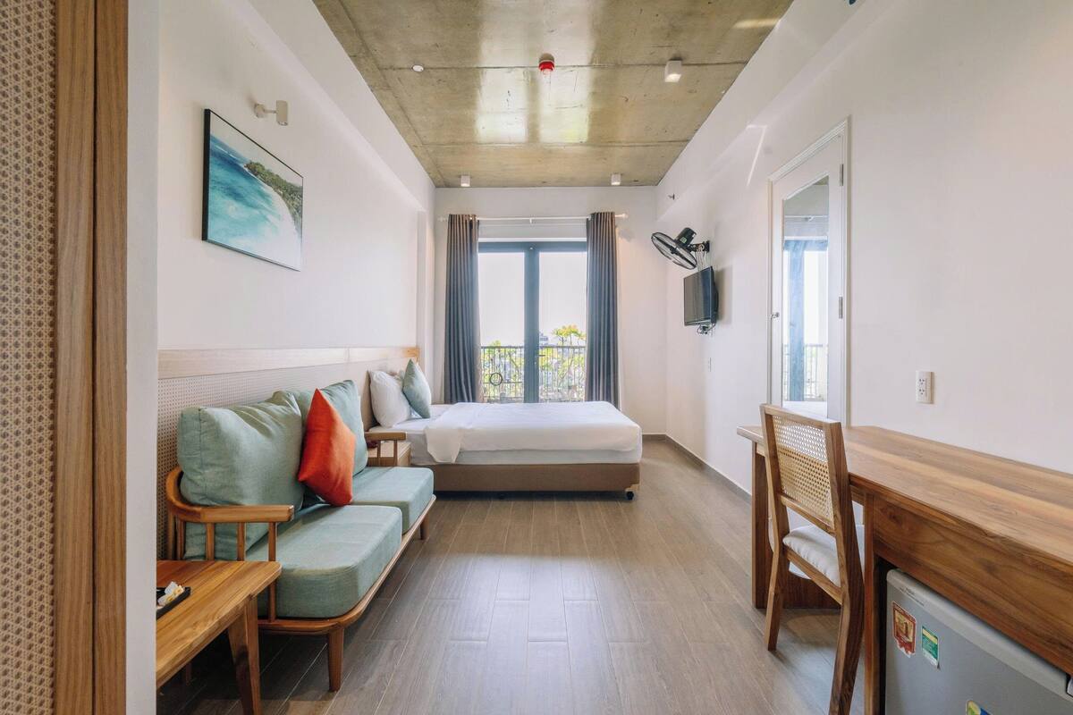 Ein Zimmer mit einem Bett, einem Holztisch, einem Sessel, einem Sofa, einem Fernseher und einem Fenster mit Vorhängen.