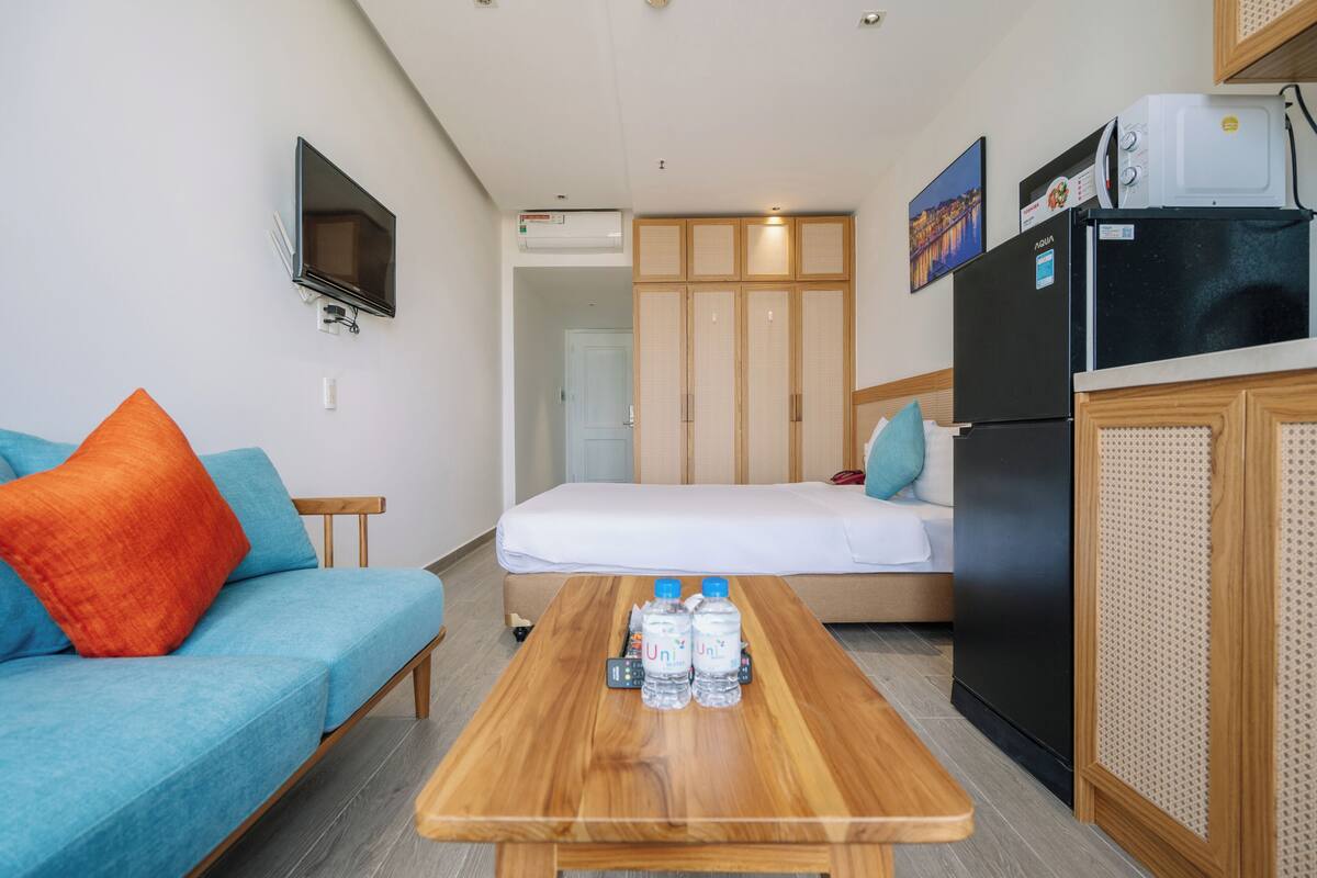 Ein kompaktes Hotelzimmer mit Sofa, Bett, Holztisch mit Wasserflaschen, einem schwarzen Kühlschrank und einem an der Wand befestigten Fernseher.