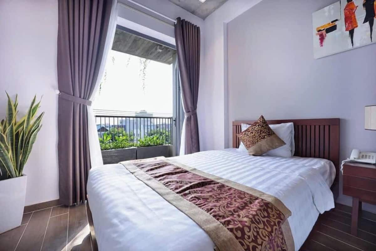 Ein Hotelzimmer mit einem großen Bett, einem Blumentopf, einem Balkon mit Vorhängen und einem Gemälde an der Wand.