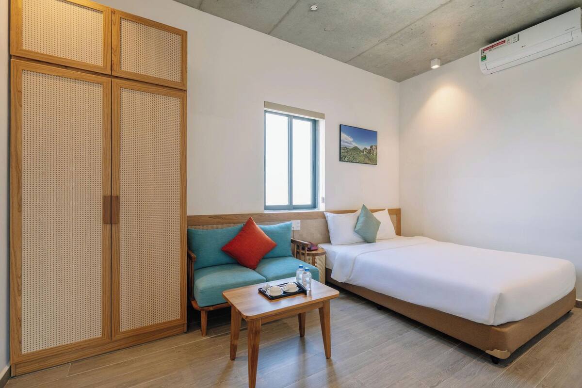 Ein Zimmer mit einem Bett, einem Schlafsofa, einem kleinen Tisch mit Gegenständen, einem Fenster und einem Bild an der Wand.
