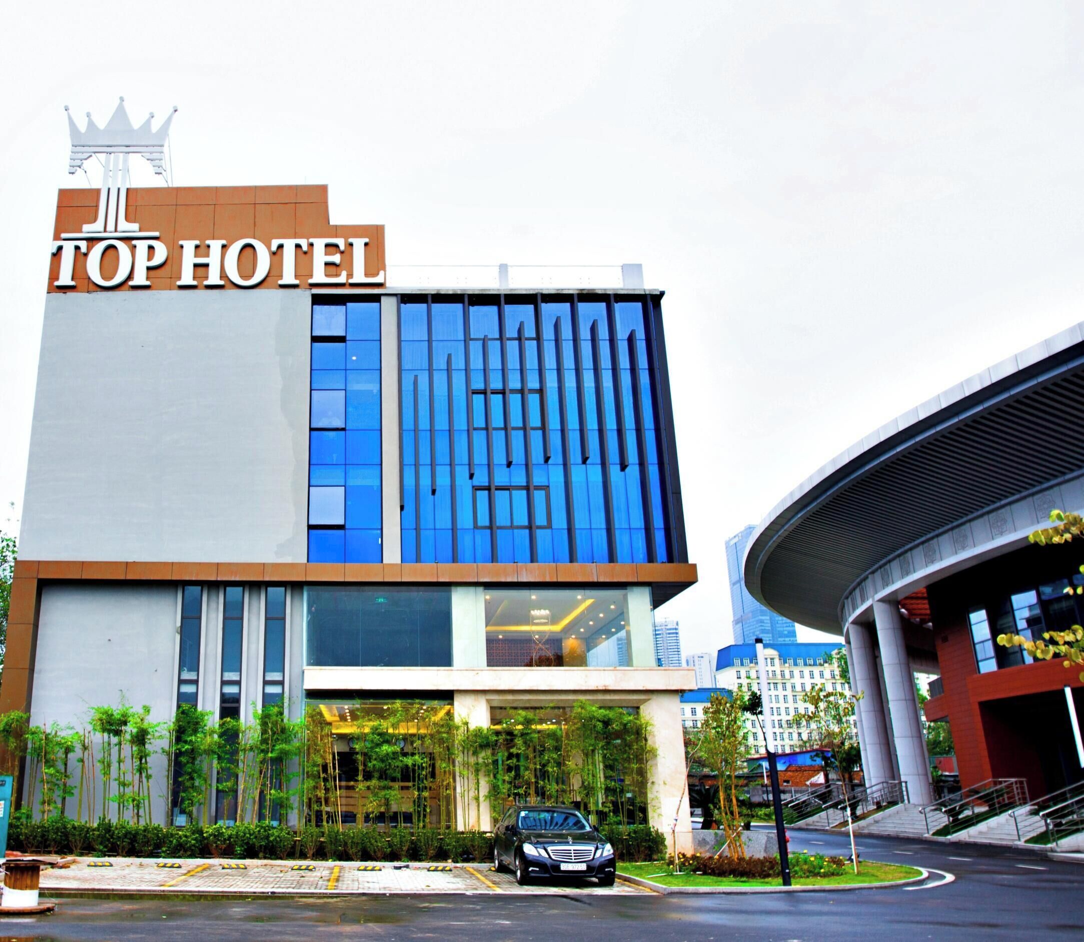 A modern hotel building with a prominent "TOP HOTEL" sign, a large glass facade, and a parking area in front.