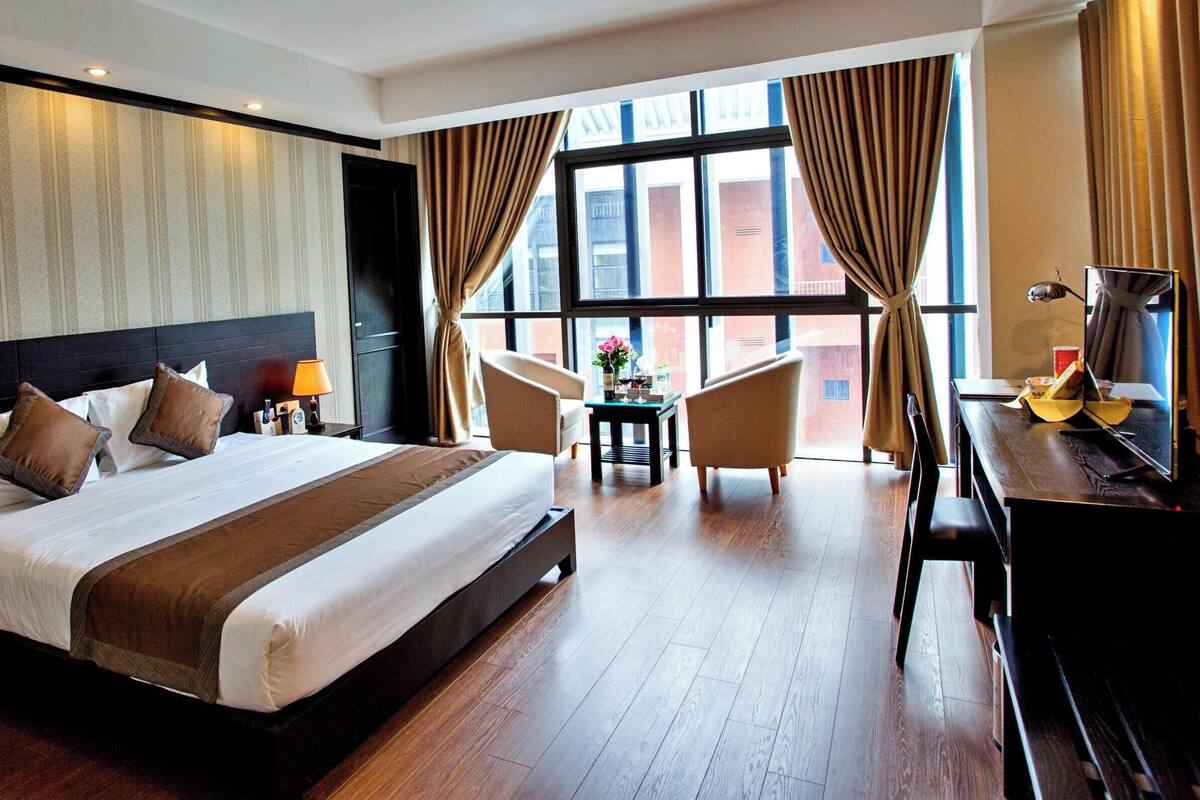 A hotel room with a large bed, a desk with a piano, a chair, a small table with flowers, and a window with curtains.
