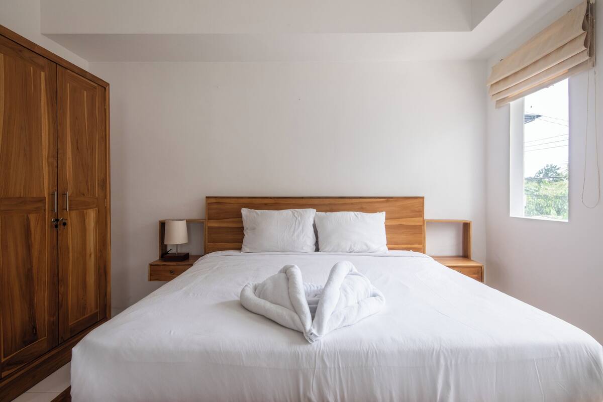 A neatly made bed with a towel in the shape of a heart, a wooden wardrobe, a bedside table with a lamp, and a window with curtains.