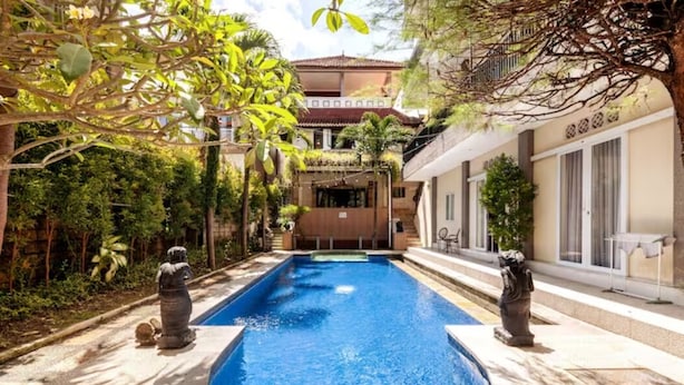 Piscina com duas estátuas de cada lado, rodeada por vegetação e um edifício com telhado telhado ao fundo.