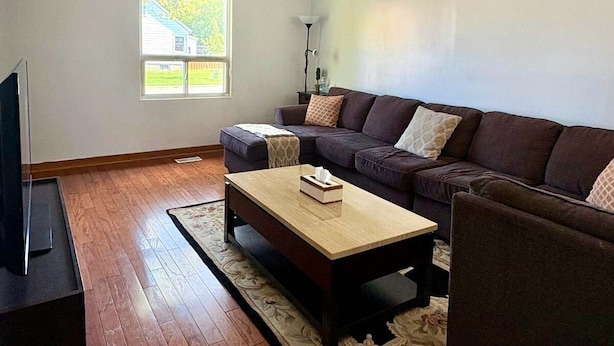 Een woonkamer met een zithoek, een salontafel, een tv-meubel en een raam met uitzicht op een huis.