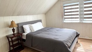 Een slaapkamer met een houten vloer, een bed met een grijze dekbedovertrek, witte kussens en een nachtkastje van hout met een lamp.