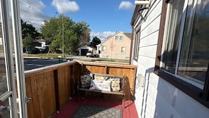 Een klein terras buiten met een houten bankje en vloerkleed, naast een gebouw met grote ramen.
