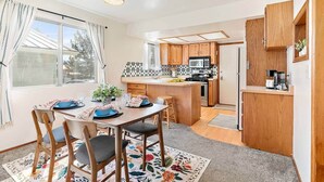 A modern kitchen with wooden cabinets, a central island, and a dining area with a table set for two. - Quaint Swiss Chalet • Steps from Main St- Sleeps 5 (Midway)