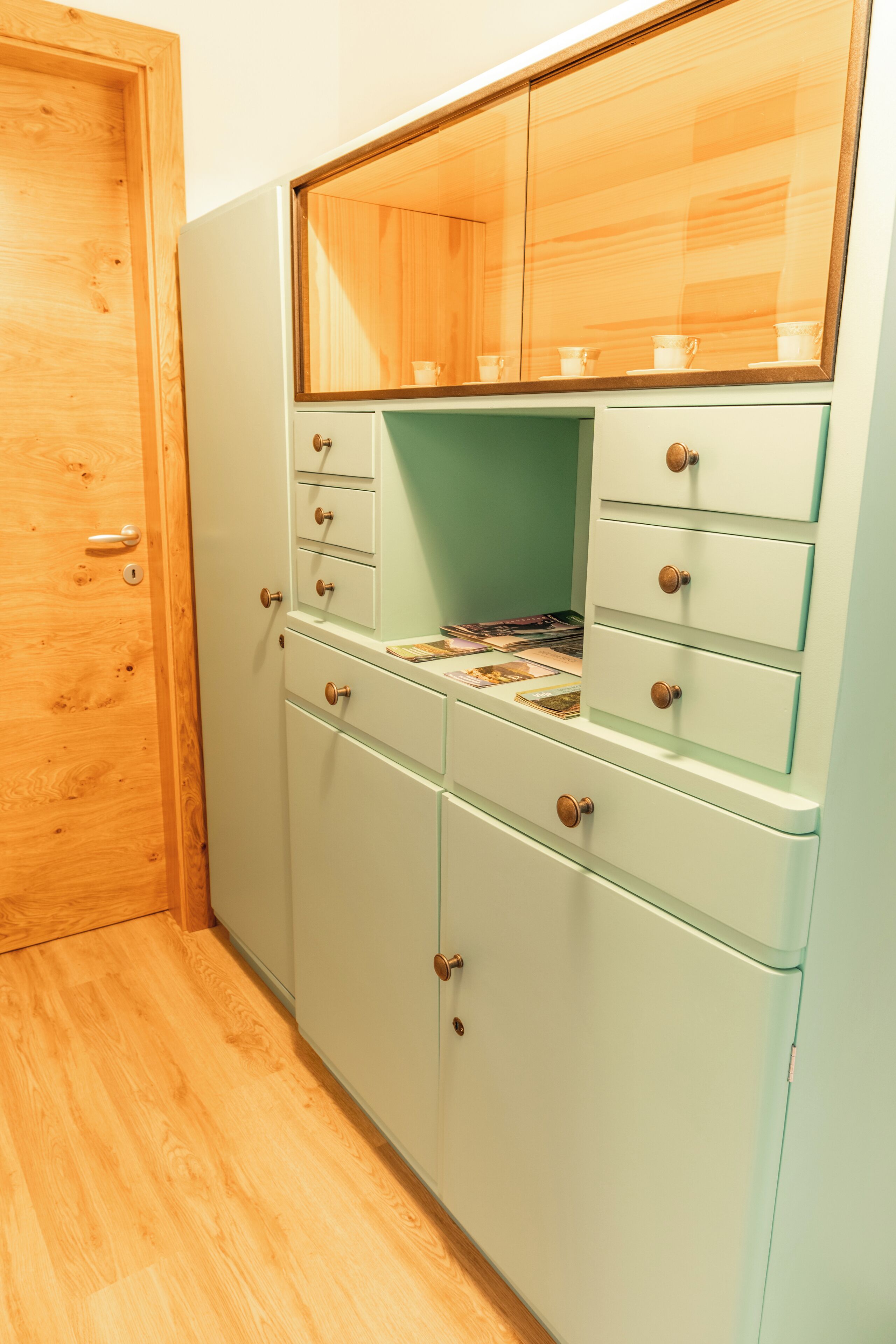 Una stanza con una porta in legno, un mobiletto verde chiaro con diversi cassetti e uno specchio sopra il mobiletto.