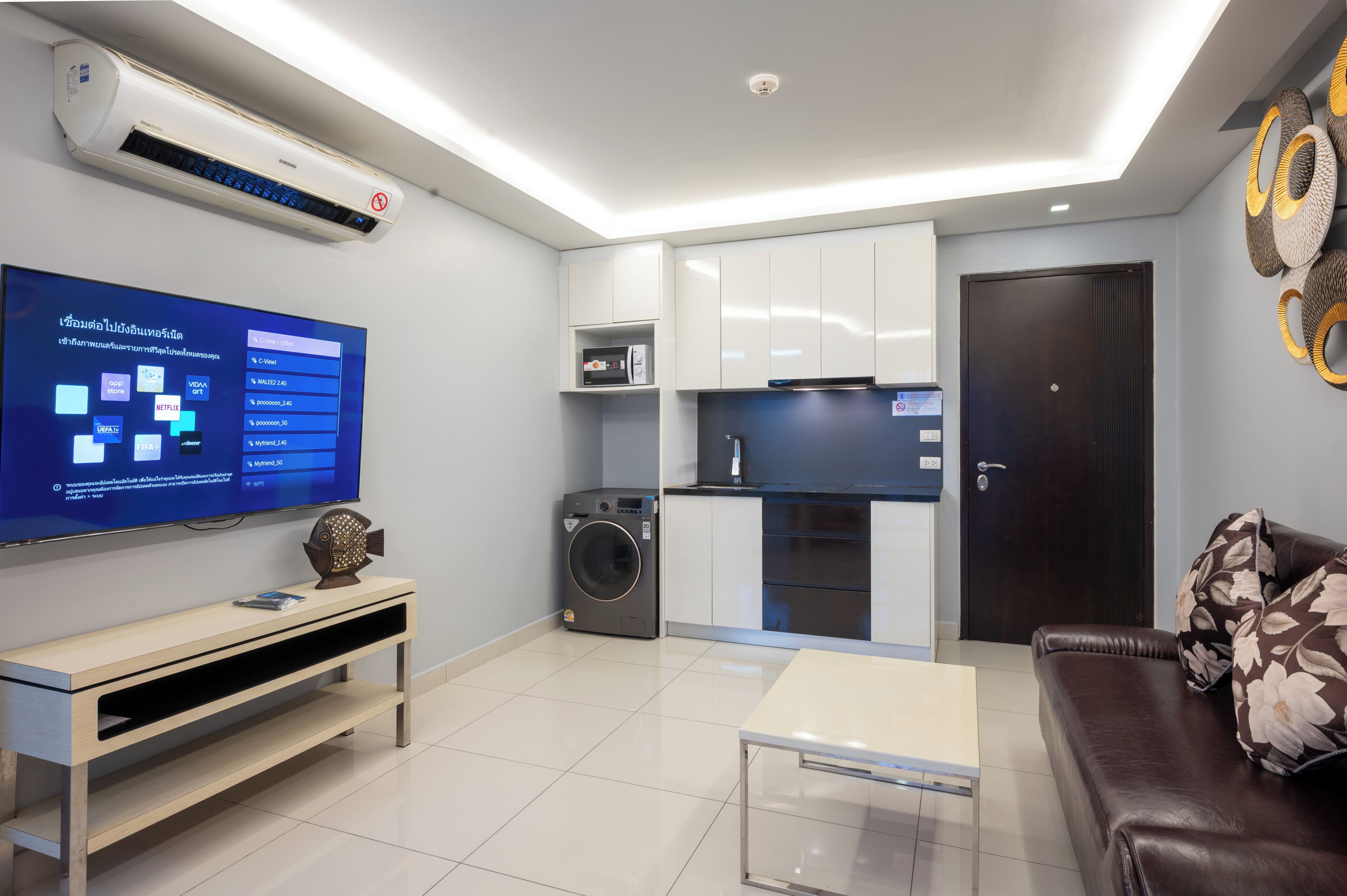 A modern living room with a flat-screen TV, a washing machine, a small kitchen area, and a dark brown sofa.