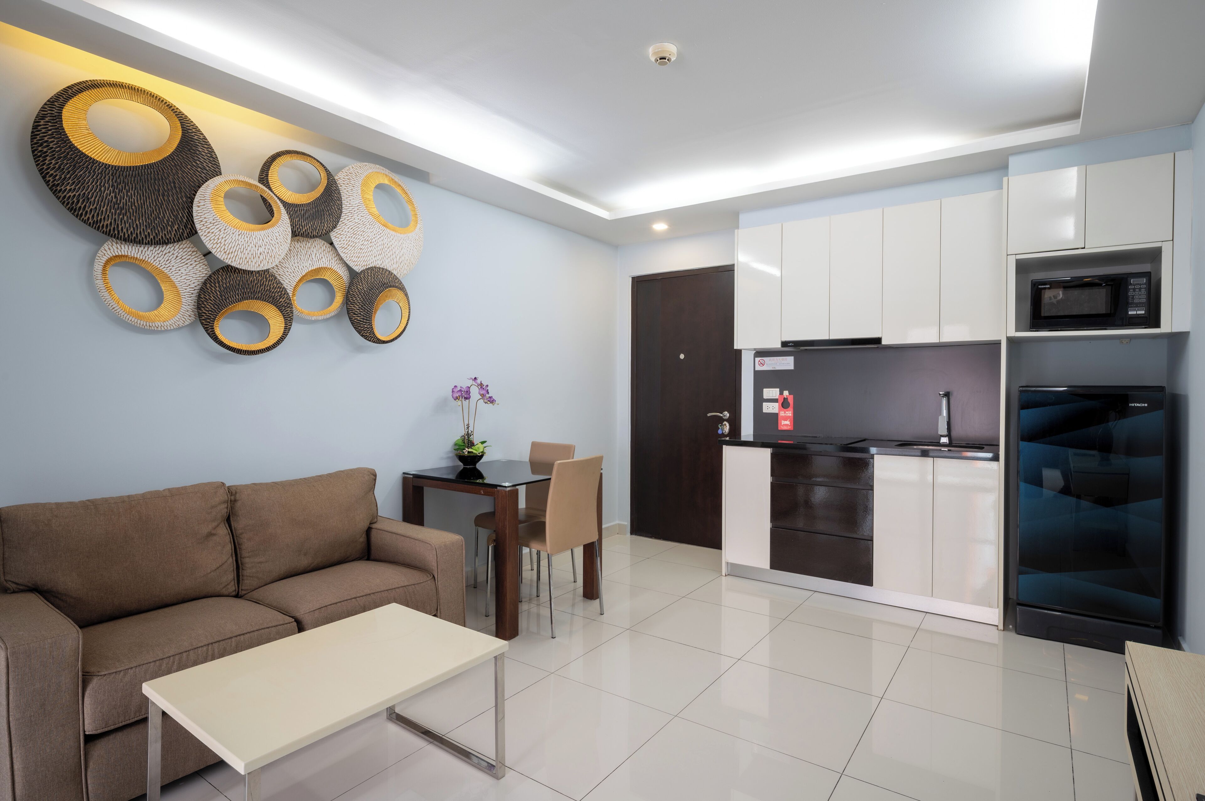 A modern living room with a sofa, coffee table, and a decorative wall piece. A kitchen with white cabinets and appliances is visible in the background.