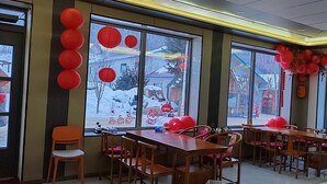 Egy étkező, melyben fából készült asztalok és székek találhatók, nagy ablakai piros díszítéssel vannak ellátva, és kilátást nyújt a havas tájra.