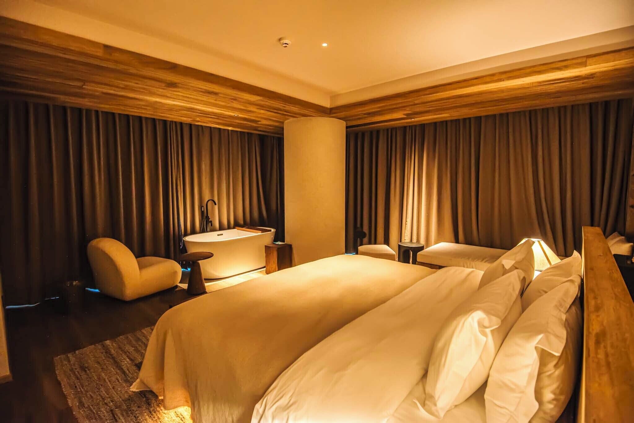 A hotel room with a large bed, a sitting area with a chair and a small table, a round mirror, and a large window with curtains.