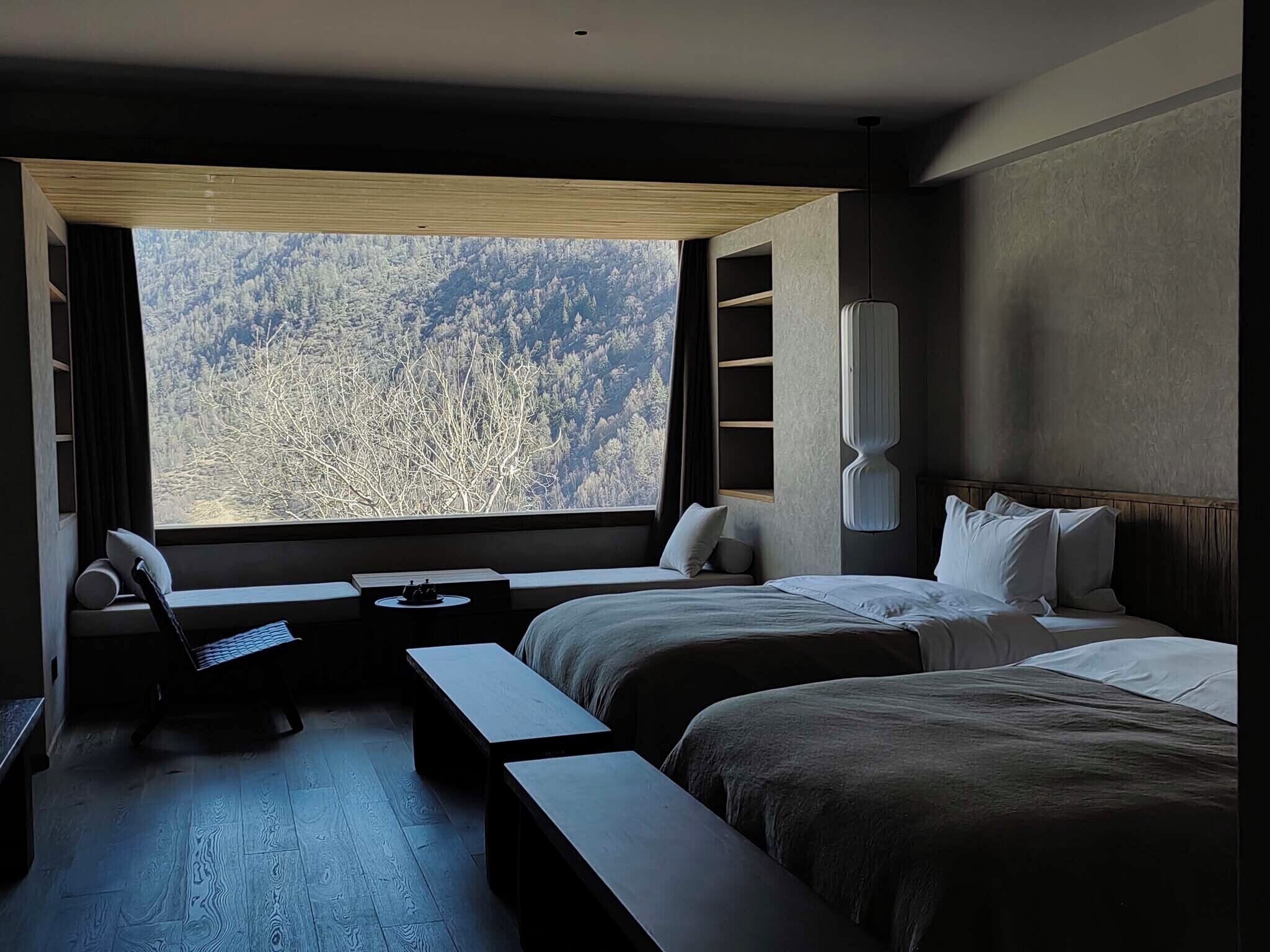 A modern hotel room with a large window offering a view of a forested mountain range. There are two beds with white linens, a small desk with a chair, and a built-in shelf unit.