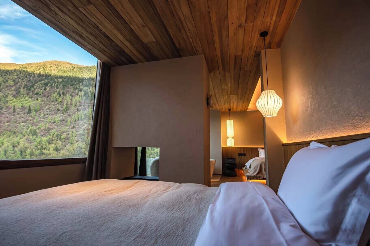 Un dormitorio con una cama grande, techo de madera, una ventana con vista a un paisaje boscoso y una mesita de noche con lámpara.