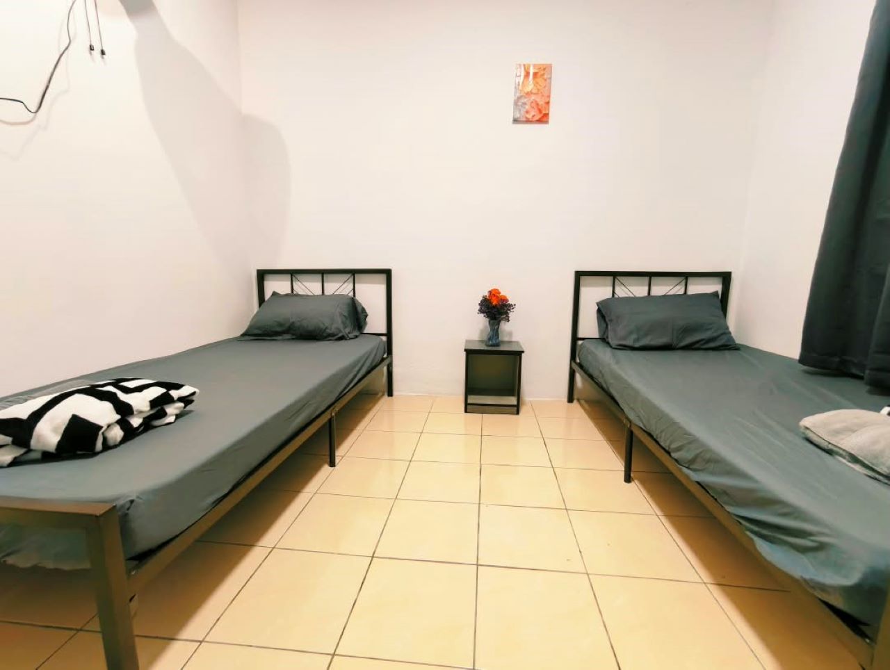 A room with two single beds, a small table with a vase, and a plain wall with a single piece of artwork.