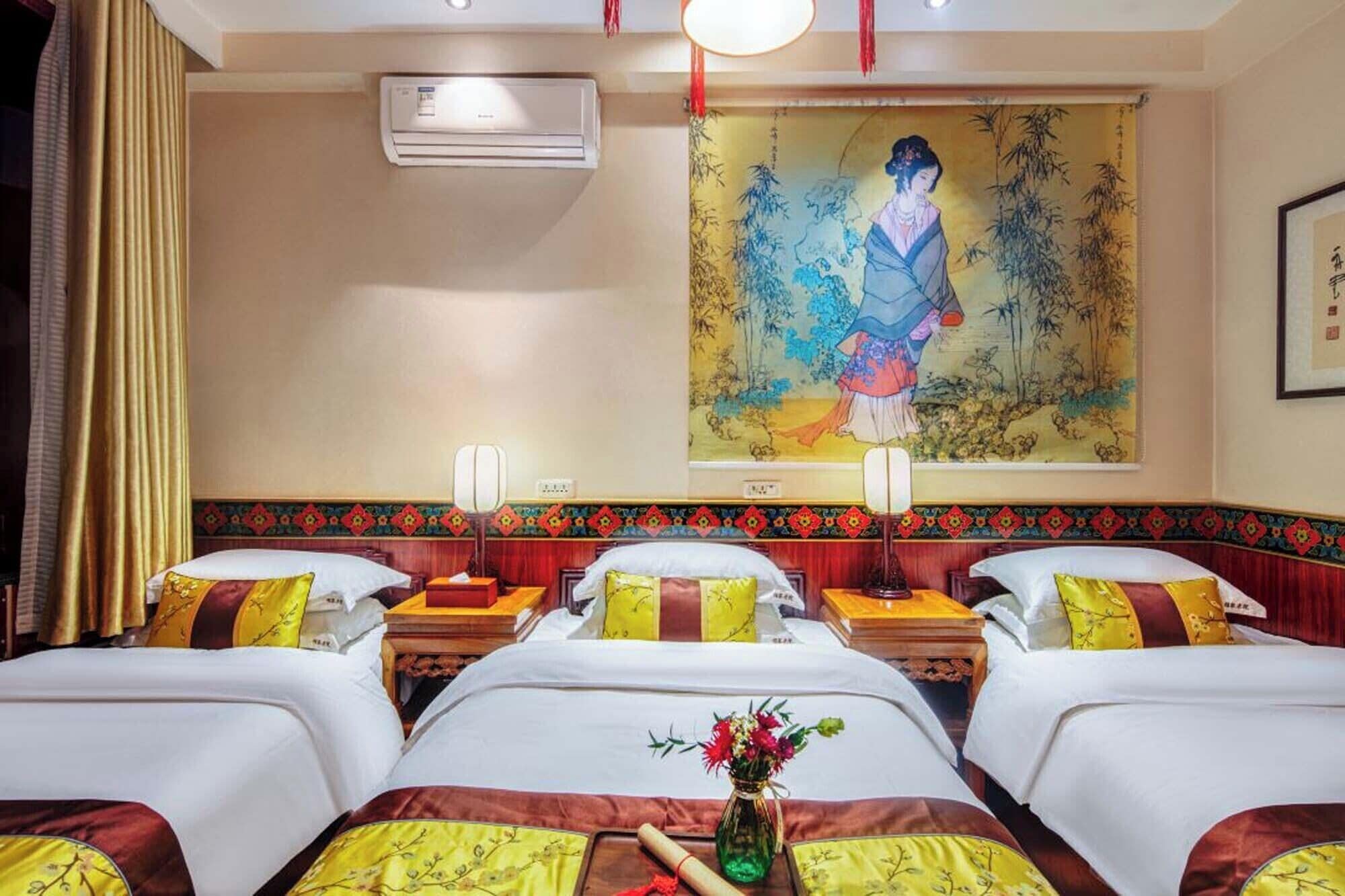 Habitación de hotel con tres camas, cada una con una colcha blanca y amarilla, una mesita con un florero y un cuadro grande de una mujer en la pared.